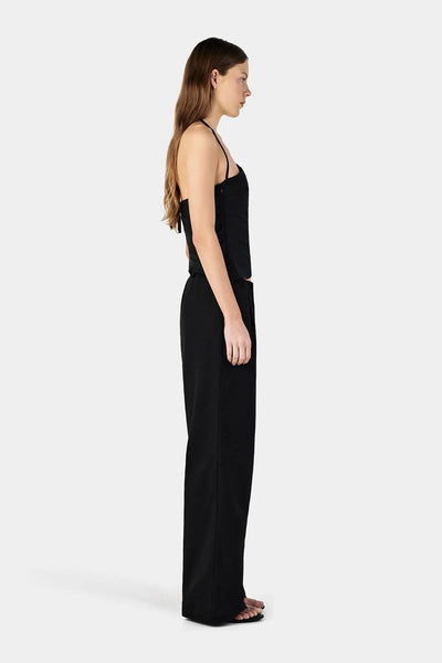 Studio side profile view of a model wearing a black asymmetrical halter top featuring a thin neck strap paired with black mid-low rise pants.