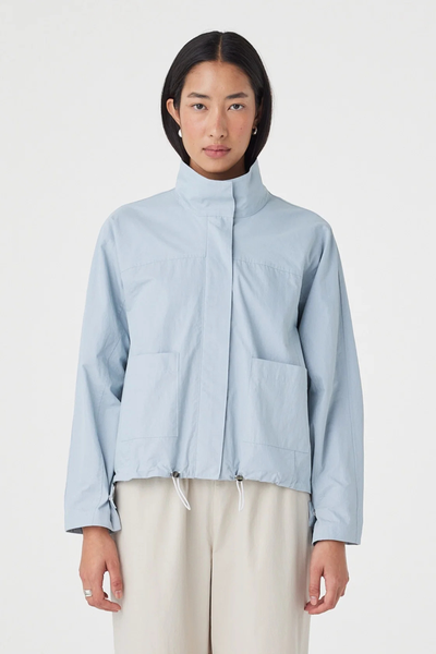 Cropped studio portrait of a model wearing the pale blue Arcaa Movement Addie Jacket zipped up with a funnel neck, showing the concealed placket, patch pockets, and adjustable hem toggles.