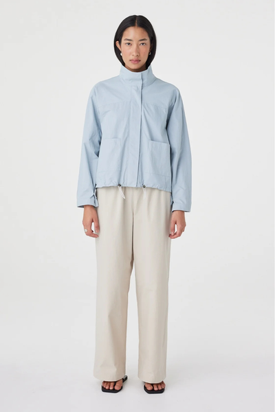 Full-length studio shot of a model in the pale blue Arcaa Movement Addie Jacket holding the jacket front, paired with relaxed beige pants.