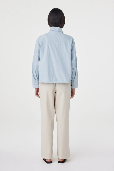 Full-length studio back view of a model wearing the pale blue Arcaa Movement Addie Jacket, detailing the clean back shoulder yoke and straight hemline.