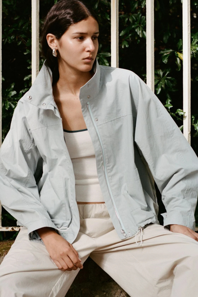 Outdoor seated shot of a model wearing the pale blue Arcaa Movement Addie Jacket unzipped over a ribbed white tank top, highlighting the snap-button funnel collar and lightweight cotton-nylon fabric.