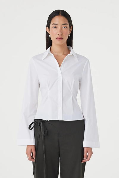Close-up front studio view of a model wearing the white 100% cotton Arcaa Movement Ari Shirt featuring front waist seams, wide cuffs, and a button-up collared neckline.