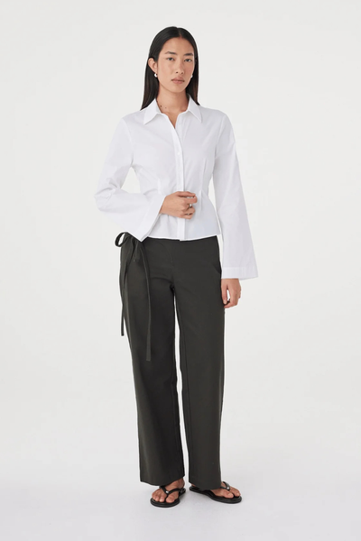 Full-length studio shot of a model posing in the white fitted Arcaa Movement Ari Shirt paired with dark olive wide-leg tie-waist trousers and black thong sandals.