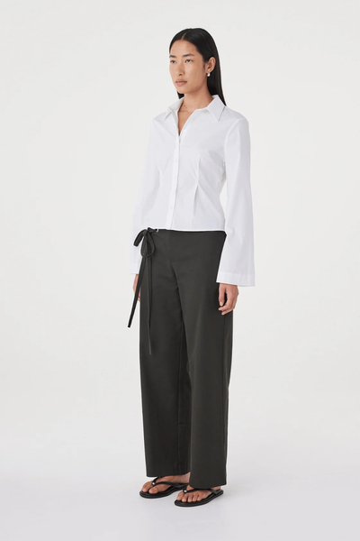 Three-quarter studio view of a model wearing the long-sleeve white Arcaa Movement Ari Shirt styled with olive green tailored trousers and black sandals.