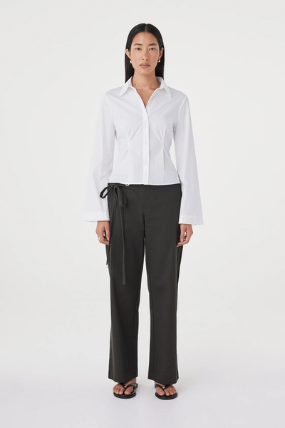 Full-length studio shot of a model posing in the white fitted Arcaa Movement Ari Shirt paired with dark olive wide-leg tie-waist trousers and black thong sandals.