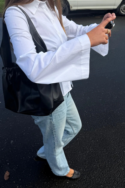 Side-angle outdoor shot of a model wearing the fitted white Arcaa Movement Ari Shirt with flared cuffs, holding a compact camera while wearing a black shoulder bag and light wash jeans.