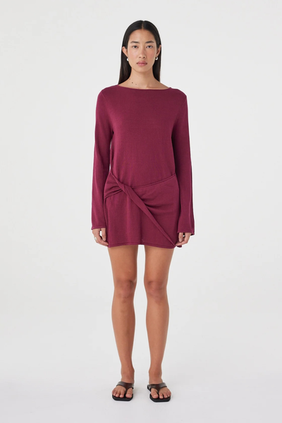Full-length studio front view of a model in the merlot Arcaa Movement Kira Mini Dress, showcasing the boat neckline, long sleeves, draped knot detail, and relaxed mini length.