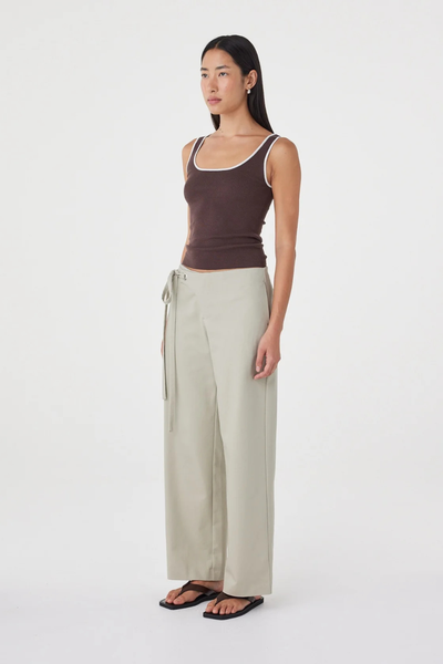 Three-quarter studio view of a model wearing the light sage Arcaa Movement Lucia Tie Pant paired with dark brown flat thong sandals and a brown tank top.