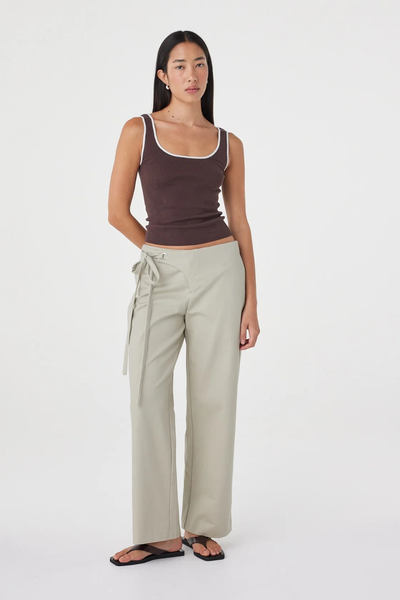 Full-length studio front shot of a model posed in the light sage Arcaa Movement Lucia Tie Pant with hands behind her back, paired with a fitted brown tank top.