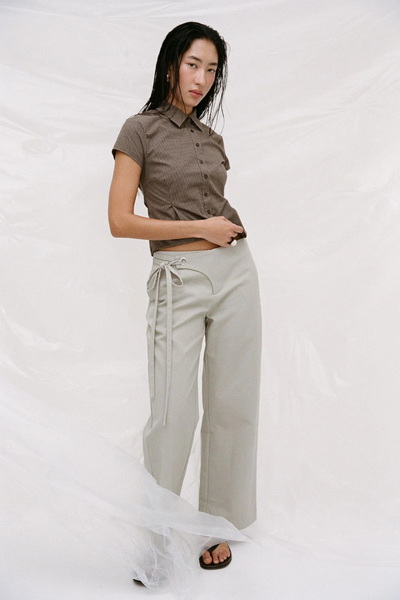 Editorial studio shot of a model wearing the light sage Arcaa Movement Lucia Tie Pant and a short-sleeve brown plaid button-up shirt behind a sheer fabric overlay.
