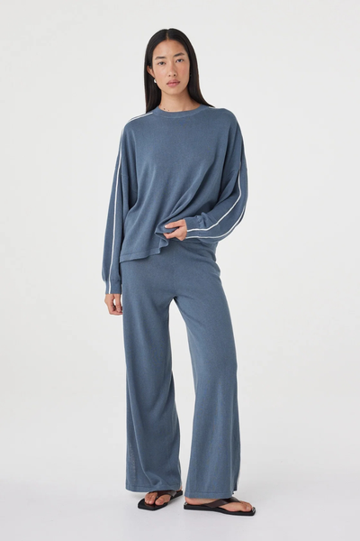 Full-length studio shot of a model holding the hem of the relaxed storm blue Arcaa Movement Pippa Long Sleeve Tee, styled with matching wide-leg knit pants.