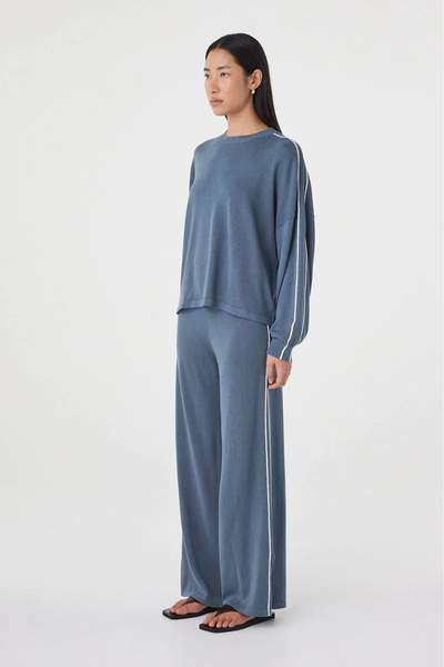 Three-quarter studio view of a model in the storm blue Arcaa Movement Pippa Pant 2.0 wide-leg knit set with cream side stripe accents, paired with dark flat thong sandals.