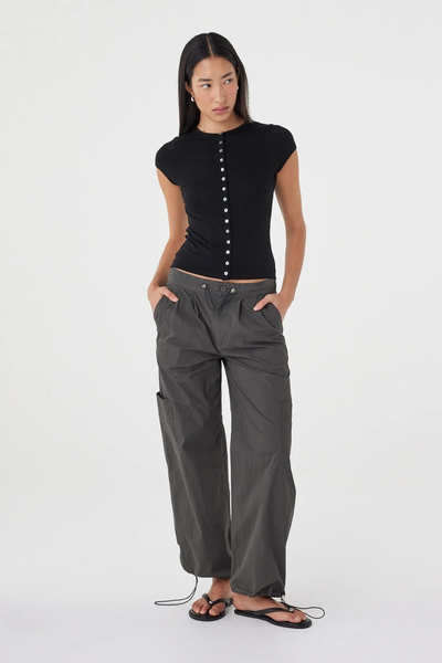 Full-length studio shot of a model wearing the black lightweight knit Arcaa Movement Robbie Top with hands in the pockets of dark charcoal wide-leg cargo pants.