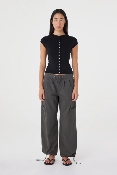 Full-length studio front view of a model wearing the black tencel-blend Arcaa Movement Robbie Top with cap sleeves, styled with relaxed dark grey utility trousers and flat sandals.