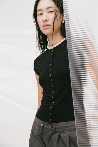Editorial close-up of a model wearing the black button-front Arcaa Movement Robbie Top with a pearl necklace, posed behind a textured glass panel.