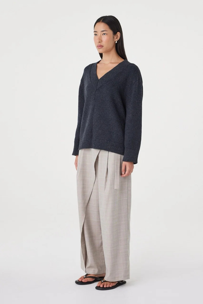 Three-quarter studio view of a model wearing the deep V-neck Arcaa Movement Zenya Sweater in dark grey marle, paired with light taupe wide-leg pants.