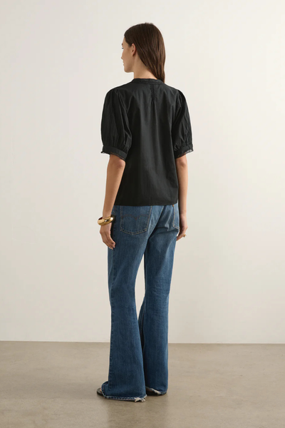 Full-length studio back-view of a model wearing the black Auguste Magdalena Blouse. Styled with classic blue flare jeans and a chunky gold bangle, the shot highlights the clean back panel construction, the relaxed fit, and the subtle lace trim bordering the short puff sleeves.