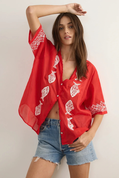 Studio lifestyle shot of a model posing with one arm raised, wearing the red oversized button-down blouse with white embroidered sardine motifs and denim shorts.