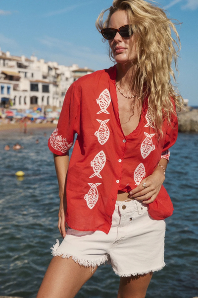 Outdoor beachside portrait of a model wearing sunglasses and the red embroidered blouse paired with white denim shorts.