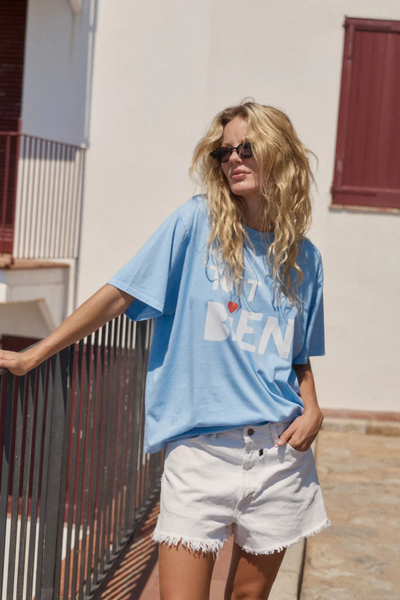 Outdoor lifestyle shot of a model with blonde hair posing against a sunny white building while wearing the sky blue slogan tee and white denim shorts.