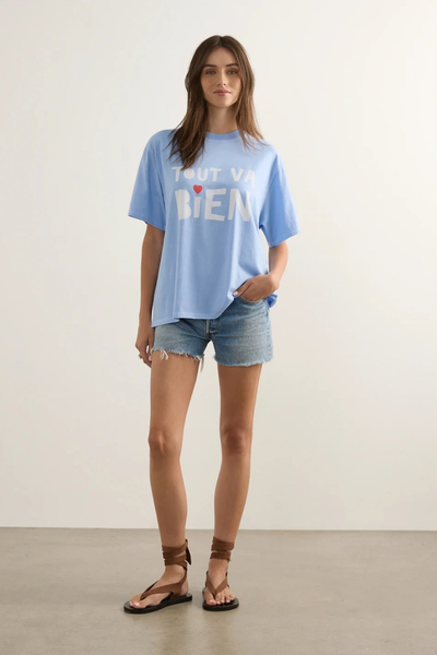 Full-length studio view of a model standing in the relaxed-fit sky blue graphic tee, denim cutoff shorts, and brown strappy sandals.