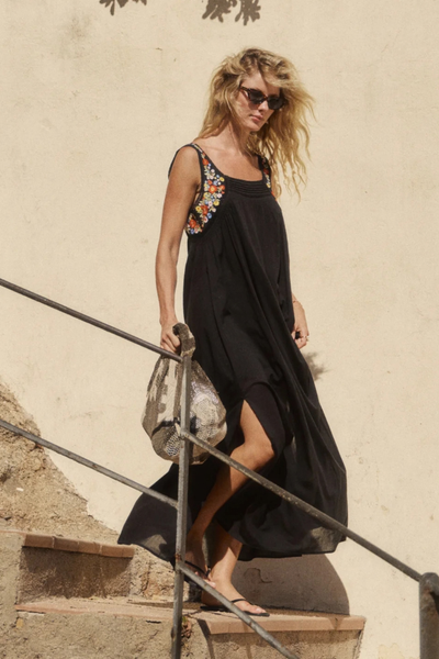 Outdoor full-length action shot of a model walking down outdoor steps while wearing the black embroidered maxi dress and carrying a patterned bag.