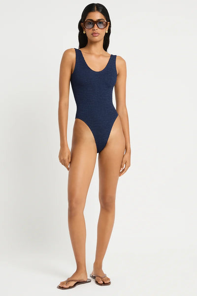 Full-length studio front view of a model wearing dark sunglasses, flat flip-flops, and the dark navy nocturne shimmer crinkle soft V-neck one-piece swimsuit.