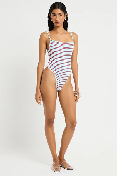 Full-length studio front view of a model wearing clear embellished flat shoes and a light blue and red micro-striped crinkle one-piece swimsuit featuring a square neckline and thin adjustable straps.