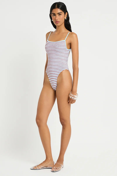 Studio three-quarter front view of a model wearing clear embellished flats and a light blue and red micro-striped square-neck one-piece swimsuit with high-cut leg lines.   