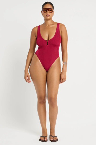 Studio front view of a curve model wearing dark sunglasses and a deep wine red shimmer cord ribbed one-piece swimsuit with center ring hardware.