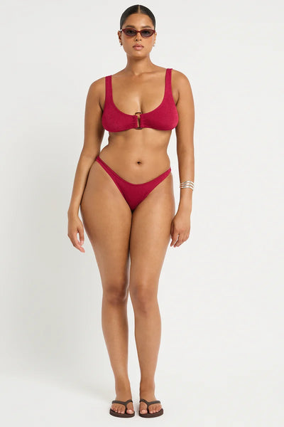 Studio front view of a curve model wearing dark sunglasses and the wine red shimmer cord ribbed Scene Brief featuring a curved waistline and thin sides, paired with a matching crop bikini top.