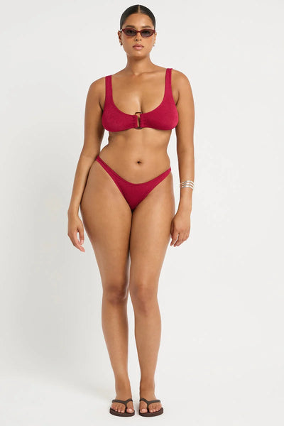 Studio front view of a curve model wearing dark sunglasses and a wine red shimmer cord ribbed scoop-neck crop bikini top featuring a center statement ring, paired with high-leg briefs.