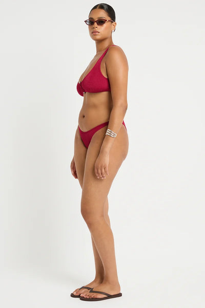 Studio side profile view of a curve model wearing the wine red shimmer cord ribbed mid-cut bikini briefs with thin side bands.