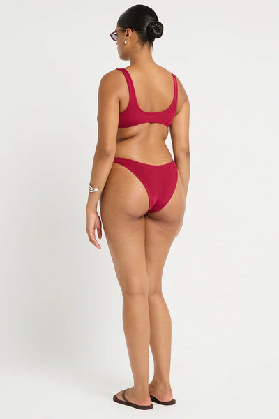 Studio rear three-quarter view of a curve model wearing dark sunglasses, flat flip-flops, and the wine red shimmer cord ribbed bikini briefs showing medium rear coverage.