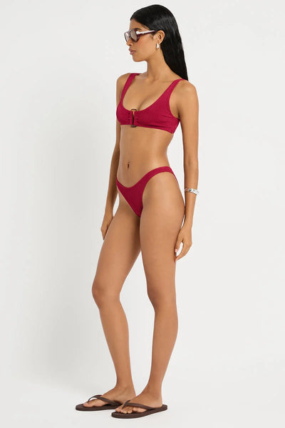 Studio three-quarter front view of a model wearing the wine red shimmer cord scoop-neck crop bikini top with a center ring detail and wide supportive straps.