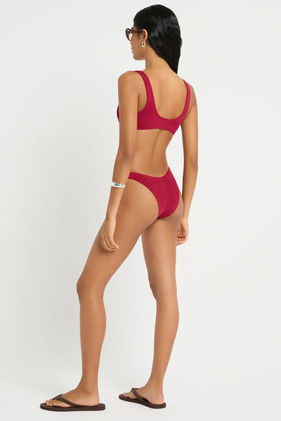 Studio rear three-quarter view of a model wearing dark sunglasses, flip-flops, and the wine red shimmer cord ribbed bikini set, highlighting the fit and coverage of the briefs.