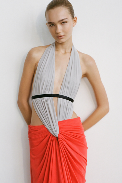 Editorial studio portrait of a model leaning against a wall wearing a dove grey halter top with a plunging neckline, black underbust strap, and flame red draped maxi skirt.