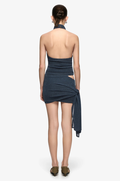 Full-length studio back view of a model with a low bun wearing a denim marle stretch-knit mini dress featuring a backless halter design, high button-closure funnel neck, right waist cutout, and a side hip sash drape.