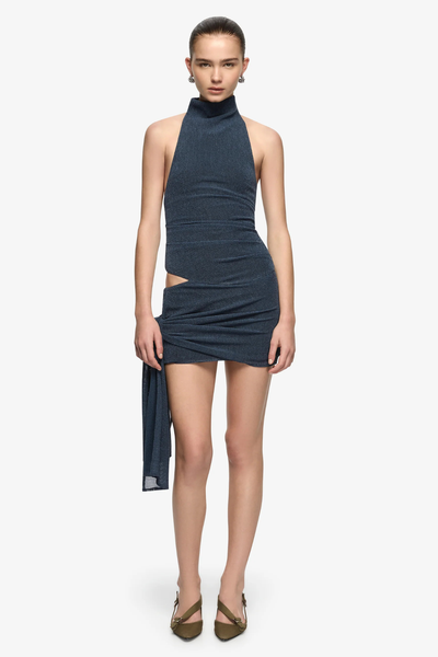 Full-length studio front view of a model wearing a high funnel-neck mini dress in denim marle stretch knit with a side hip cutout and a draped side tie feature.