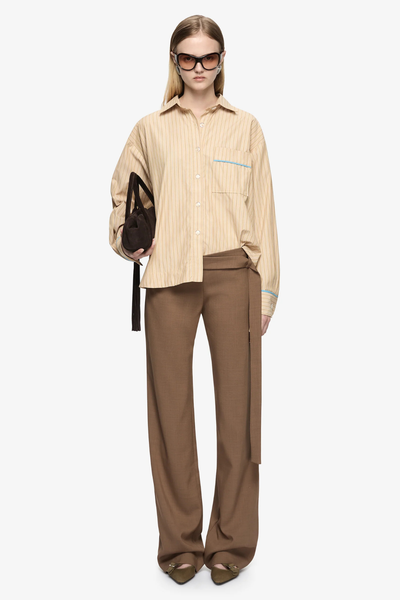 Full-body front studio view of a model wearing the Bec + Bridge Zanda Shirt in Tan Pinstripe/Azure Blue with brown trousers, sunglasses, and a handbag.