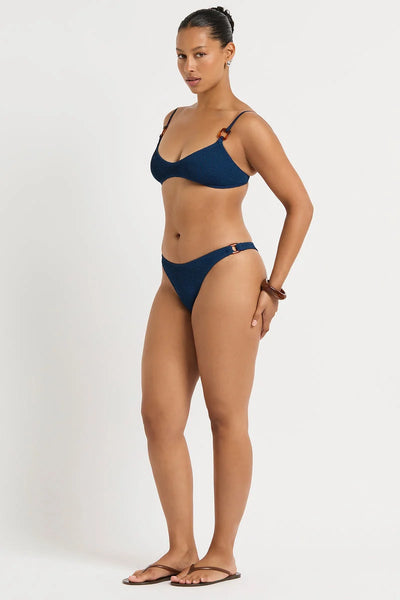 Side three-quarter standing view of model Yaz wearing the Bond-Eye Ring Scene Brief in dark blue Marine Recycled crinkle fabric, pulled high on the hip.