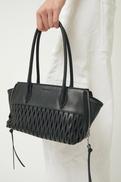 Cropped studio shot of a model carrying the black Brie Leon Faye Mini Tote in recycled leather by its dual shoulder straps, showing the laser-cut net pattern and side zip extensions against white trousers.