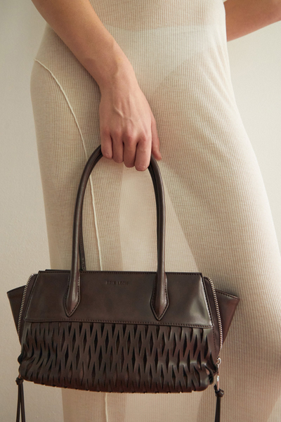 Cropped close-up shot of a model holding the dark brown Brie Leon Faye Mini Tote in ganache recycled leather by its dual shoulder straps, highlighting the laser-cut net detailing against a cream ribbed outfit.