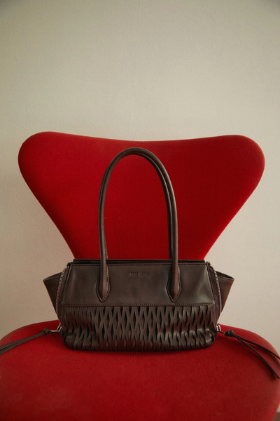 Lifestyle product photo of the dark brown Brie Leon Faye Mini Tote resting upright on a bold red modern chair against a neutral off-white wall.