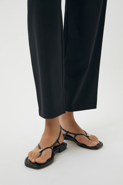 Close-up product view of a person wearing the black Halle flat sandals paired with dark trousers, showcasing the slingback strap and toe-thong design.