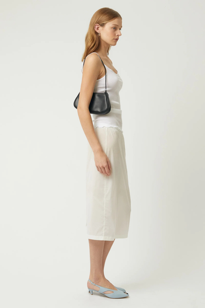 Full-length side view of a blonde model wearing the Brie Leon Iris Mini Bag in black over her shoulder with a white ribbed singlet top, midi skirt, and light blue kitten heels.