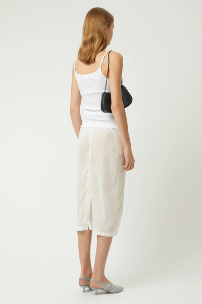 Full-length back view of a blonde model wearing the Brie Leon Iris Mini Bag in black tucked under her arm, paired with a white tank top, midi skirt, and blue heels.
