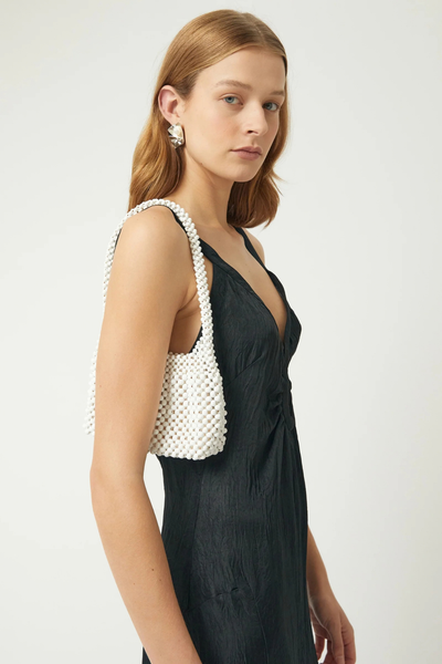 Studio side-profile portrait of a model wearing a black slip dress and earrings while carrying the oat-colored lotus wood beaded mini bag over her shoulder.