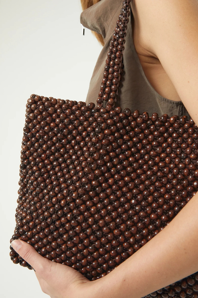 Close-up studio shot of a model holding the handwoven dark chocolate lotus wood beaded tote bag against her side.