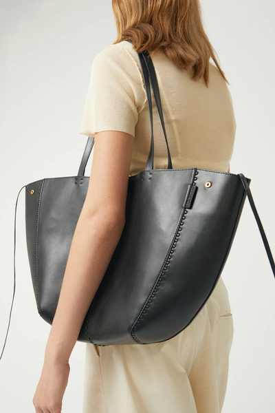 Cropped three-quarter view of a model styled in a cream short-sleeve top and cream trousers carrying the black Brie Leon Reese Market Tote in recycled leather over her shoulder in its open, wide tote configuration.